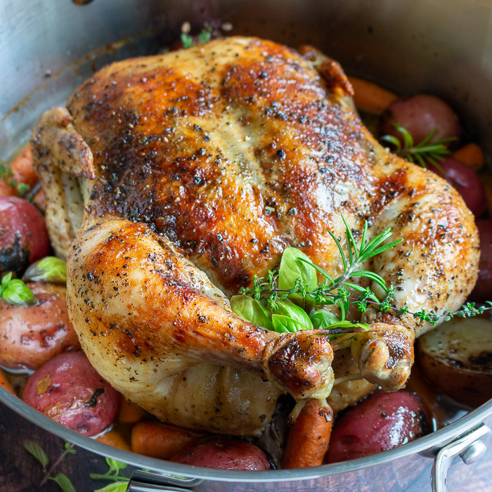 Perfectly roasted whole chicken with crispy skin, fresh herbs surrounded by cooked potatoes and carrots. 