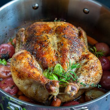 Perfectly roasted whole chicken with crispy skin, fresh herbs surrounded by cooked potatoes and carrots.