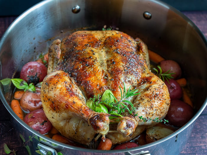 Perfectly roasted whole chicken with crispy skin, fresh herbs surrounded by cooked potatoes and carrots.