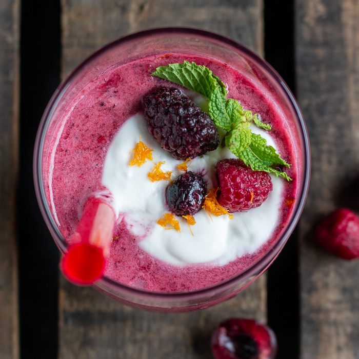 Top down shot. Berry cherry Smoothie Blast topped with a little dollop of homemade coconut whipped cream, fresh berries, mint and orange zest in a clear glass with a red straw. 