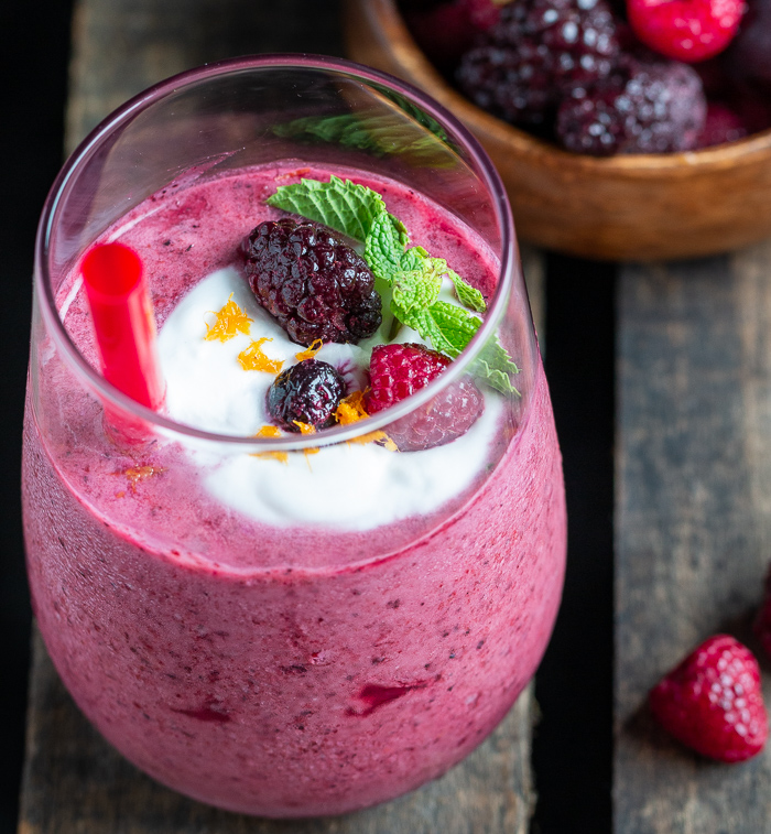 Berry cherry Smoothie Blast topped with a little dollop of homemade coconut whipped cream, fresh berries, mint and orange zest in a clear glass with a red straw. 