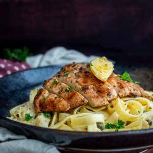 Grilled chicken slices on top of a lemon, caper, artichoke pasta with an artichoke on top and garnished with parsley