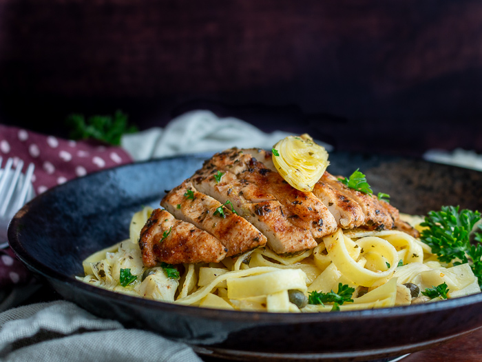 Grilled chicken slices on top of a lemon, caper, artichoke pasta with an artichoke on top and garnished with parsley