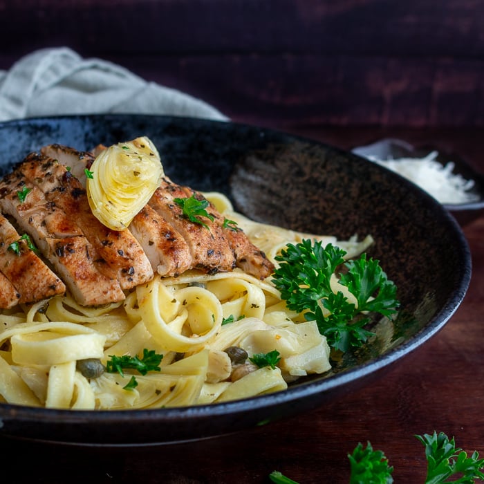 Grilled chicken slices on top of a lemon, caper, artichoke pasta with an artichoke on top and garnished with parsley on a black plate