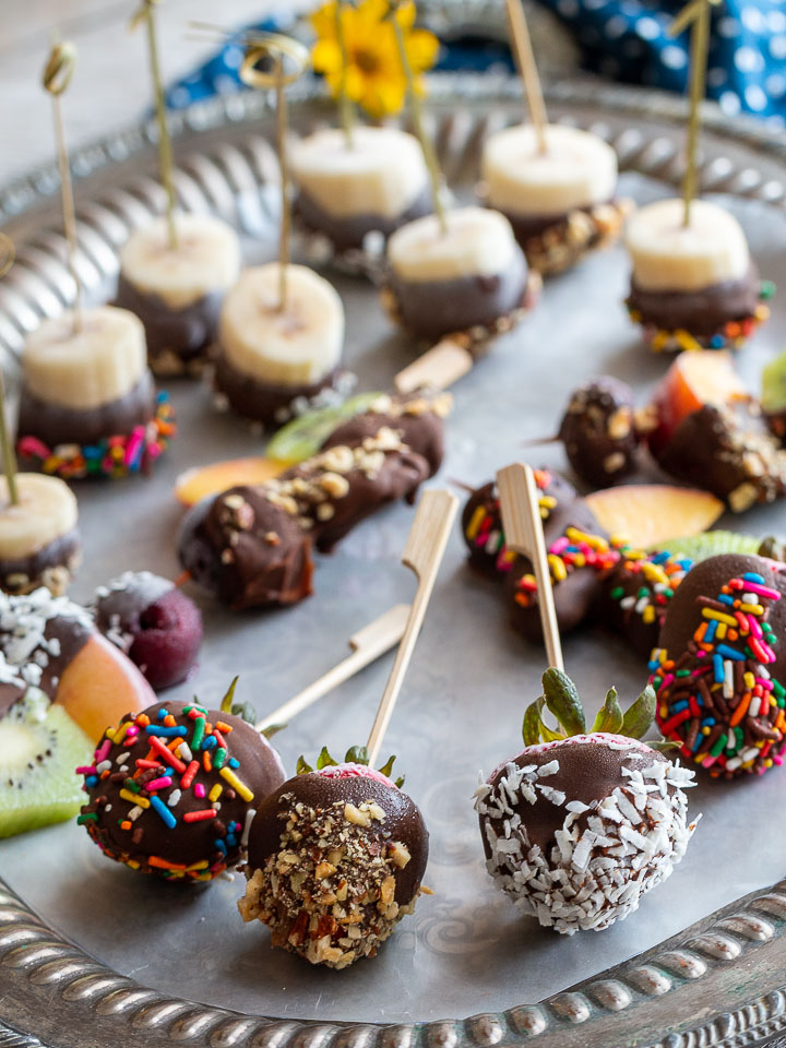 Platter of chocolate covered fruit with assorted toppings on a lined silver platter.