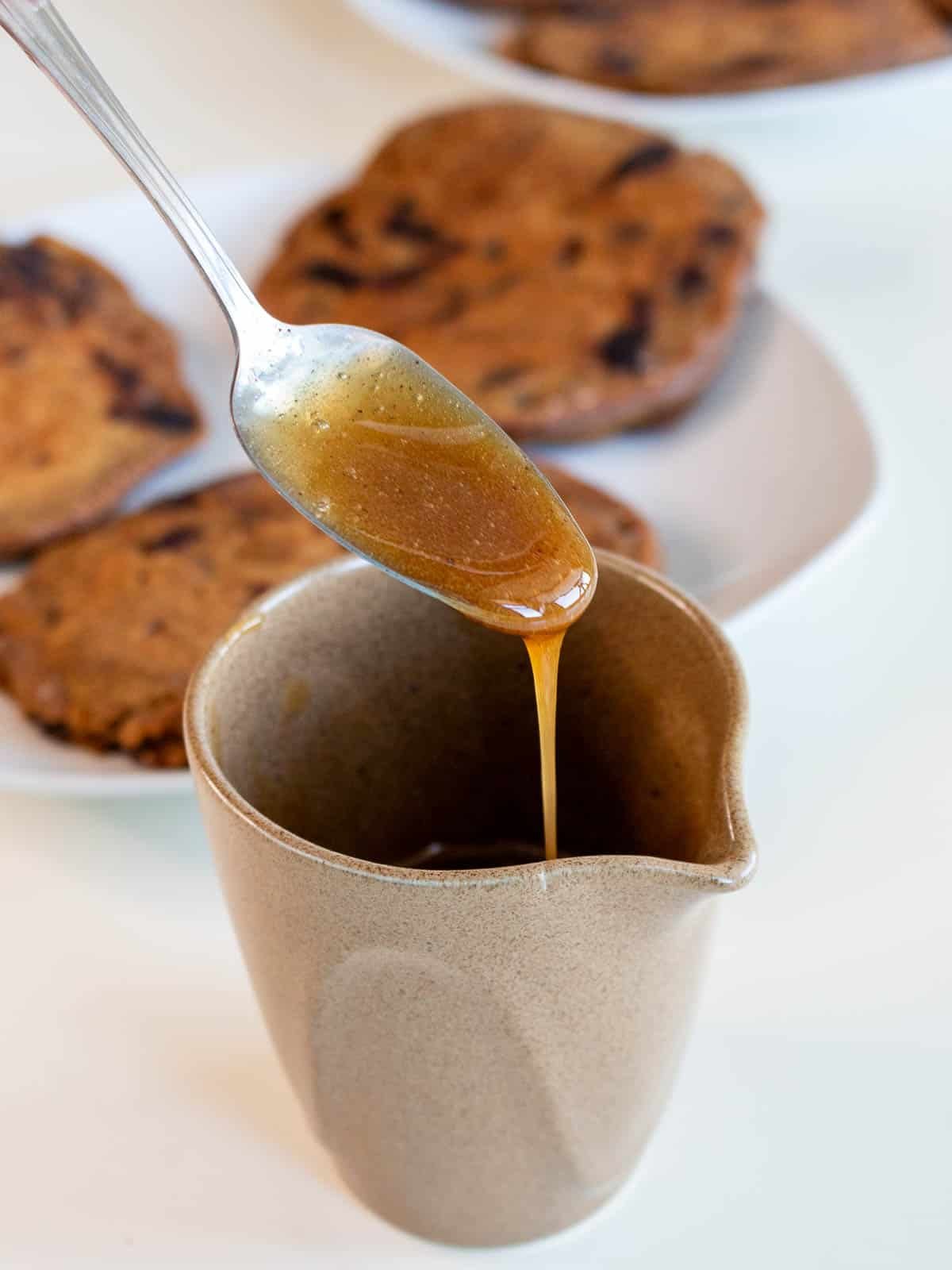 Spoonful of maple coffee syrup drizzling into a beige pouring container. 