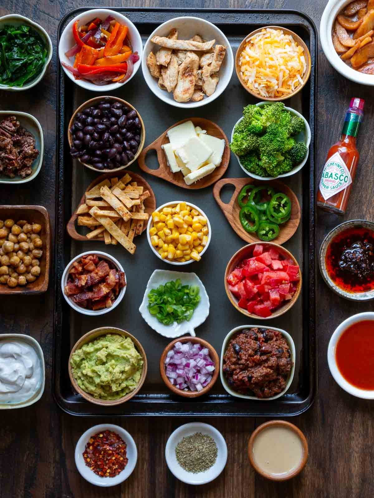 25 plus baked potato toppings chopped up and ready to go in little dishes in and around a baking tray. 