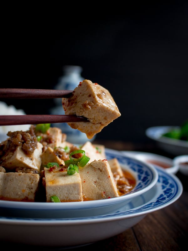 Chili covered tofu cube held by wooden chopsticks.