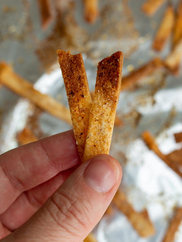 Hand holding up over the baking sheet 2 perfectly crispy baked tortilla strips. 