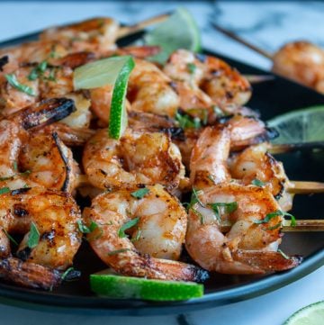 Firecracker grilled shrimp just off the grill garlished with lime and cilantro on a black plate and marble table.