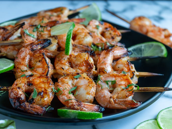 Firecracker grilled shrimp just off the grill garlished with lime and cilantro on a black plate and marble table.