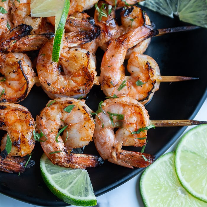 Firecracker grilled shrimp on skewers just off the grill garlished with lime and cilantro on a black plate and marble table.