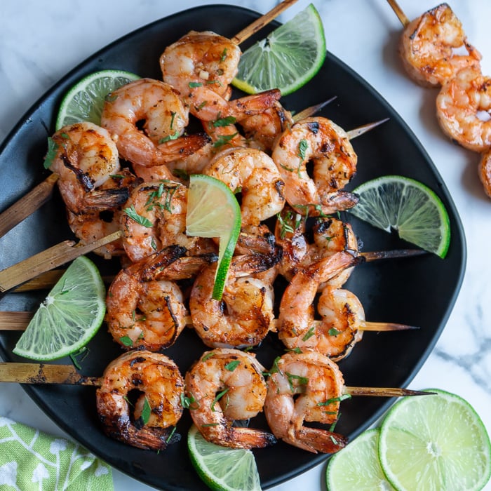 Firecracker grilled shrimp just off the grill garlished with lime and cilantro on a black plate and marble table. All criss crossed on the plate.