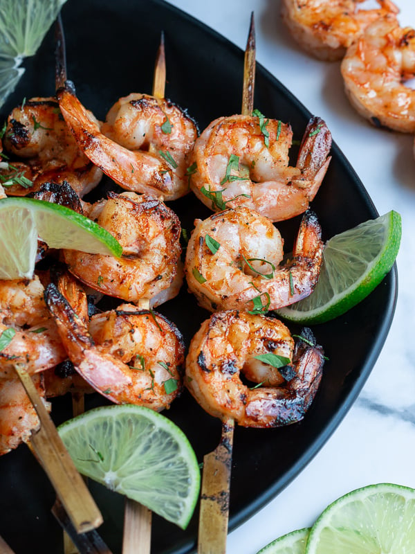 Firecracker grilled shrimp just off the grill garlished with lime and cilantro on a black plate and marble table.