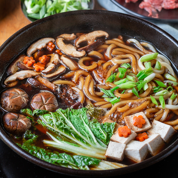 Pot of cooking sukiyaki on the table with ready to eat beef, mushrooms, udon, tofu and napa cabbage decorated with carrot flower slices. 