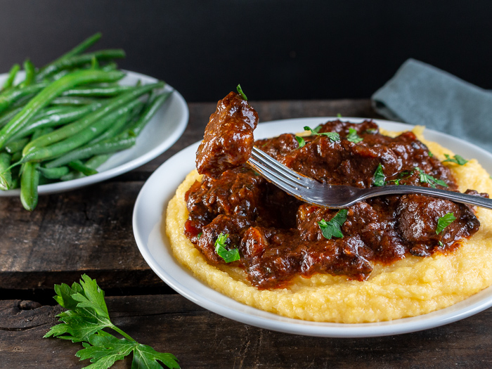 The bite shot of a juicy melt in your mouth piece of Tuscan beef over a bed of creamy polenta and a side of green beans.