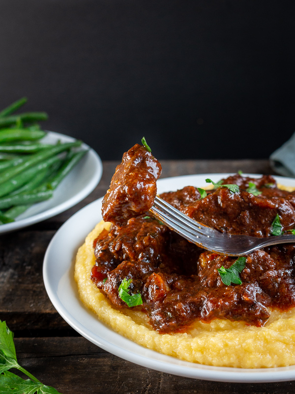 The bite shot of a juicy melt in your mouth piece of Tuscan beef over a bed of creamy polenta and a side of green beans.