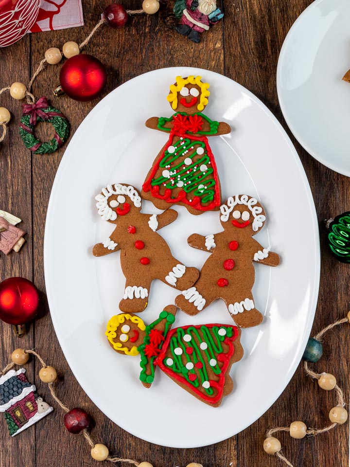 4 decorated gingerbread men and women on a white plate with holiday decorations around the plate.