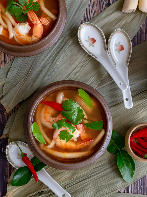 Two Tom yum goong soup bowls laid on top of bamboo leaves with fresh chilis, kaffir lime leaves and lemon grass around. 