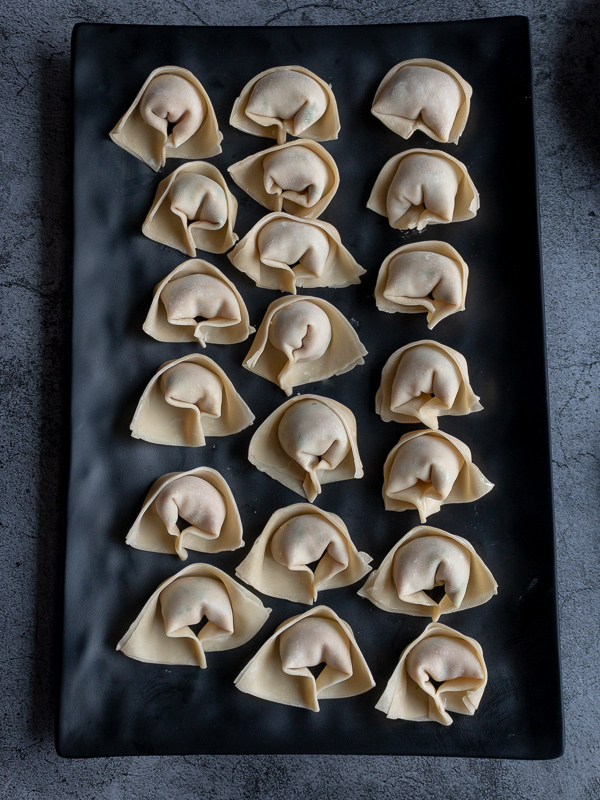 Twenty uncooked wontons all lined up on a rectangle black plate. They look like little nun caps or old fashion nurse caps.