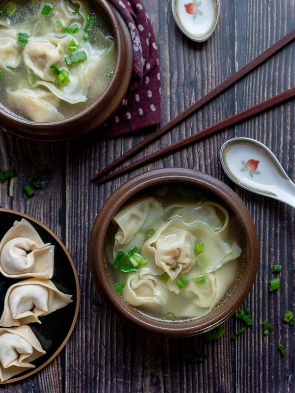 Two bowls of wontons soup, prepared wontons, chopsticks and a spoon, ready to eat.