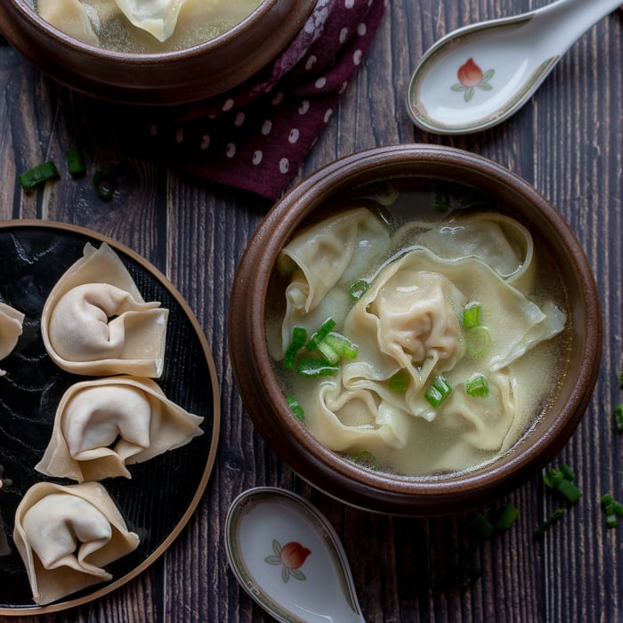 Wonton soup in a brown bowl with a plate of uncooked wontons and 2 spoons. 