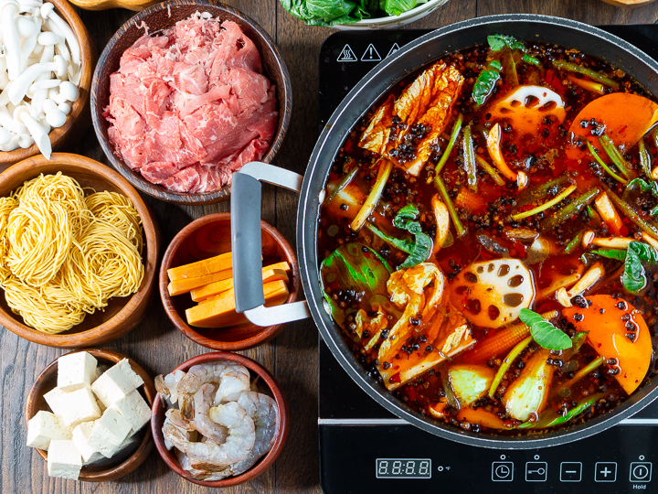 Spicy Sichuan Hot pot broth and vegetables in an electric cook top with items ready to cook in wooden bowls around the burner.