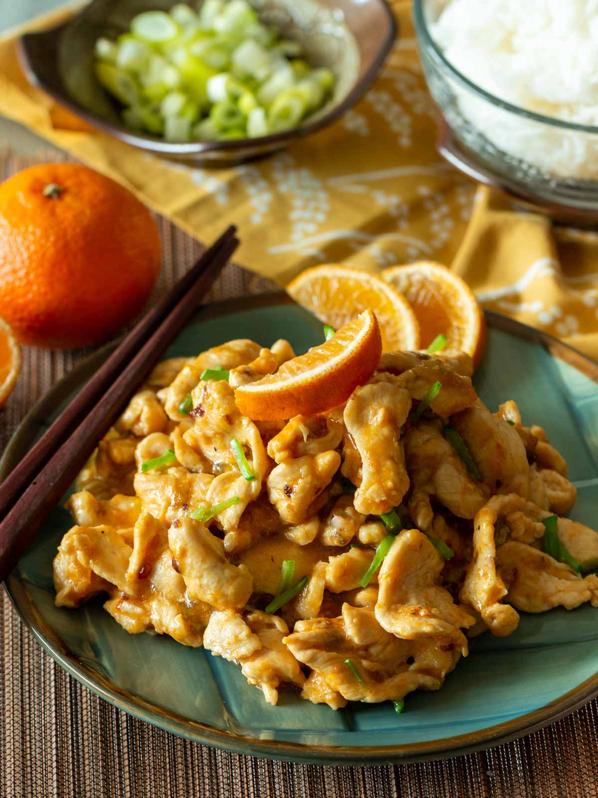 Hot and spicy mandarin orange chicken on a blue plate with white rice on the saide and garnished with oranges and green onions.