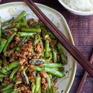 Stir fried dry fried green beans and minced pork in a Chinese bowl with wooden chop sticks and a side of rice.