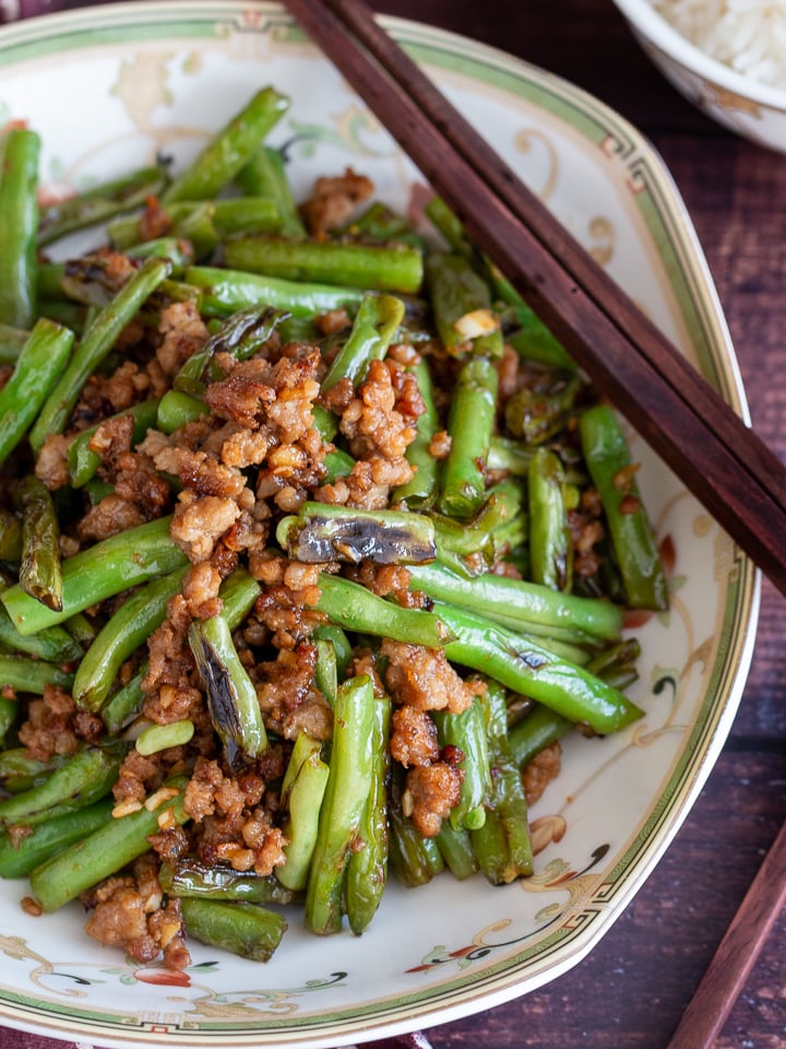 Stir fried dry fried green beans and minced pork in a Chinese bowl with wooden chop sticks and a side of rice.