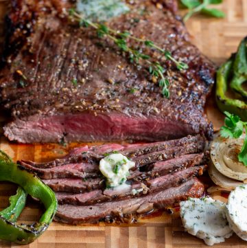 Grilled flank steak cut into thin slices on a cutting board and garnished with herb butter and fresh herbs.