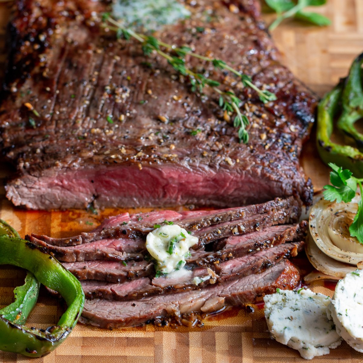 Grilled flank steak cut into thin slices on a cutting board and garnished with herb butter and fresh herbs.