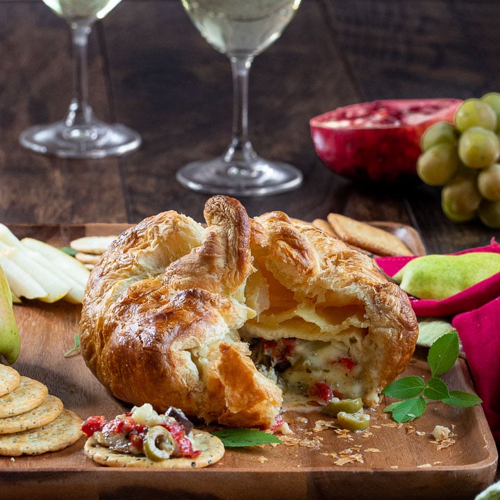 Savory Baked Brie in Puff Pastry on a cheese board cut open to show the melty cheese and Mediterranean ingredients inside. 