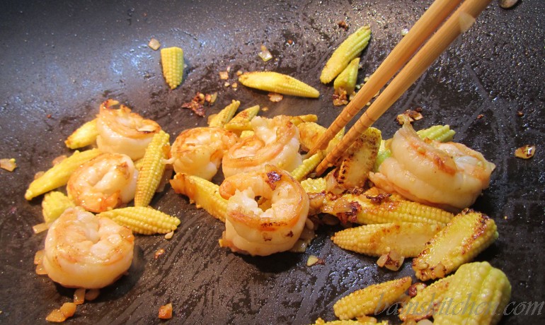 Shrimp and baby corn getting cooked in the wok with cooking chopsticks.