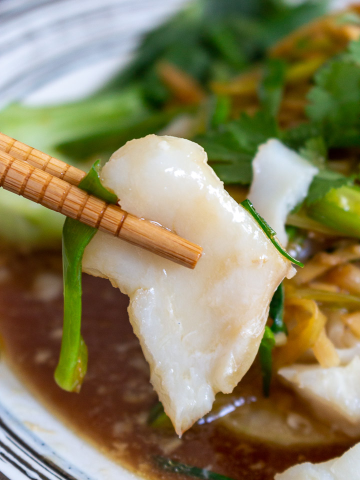 Bite shot of perfectly teamed cod held with chop sticks.