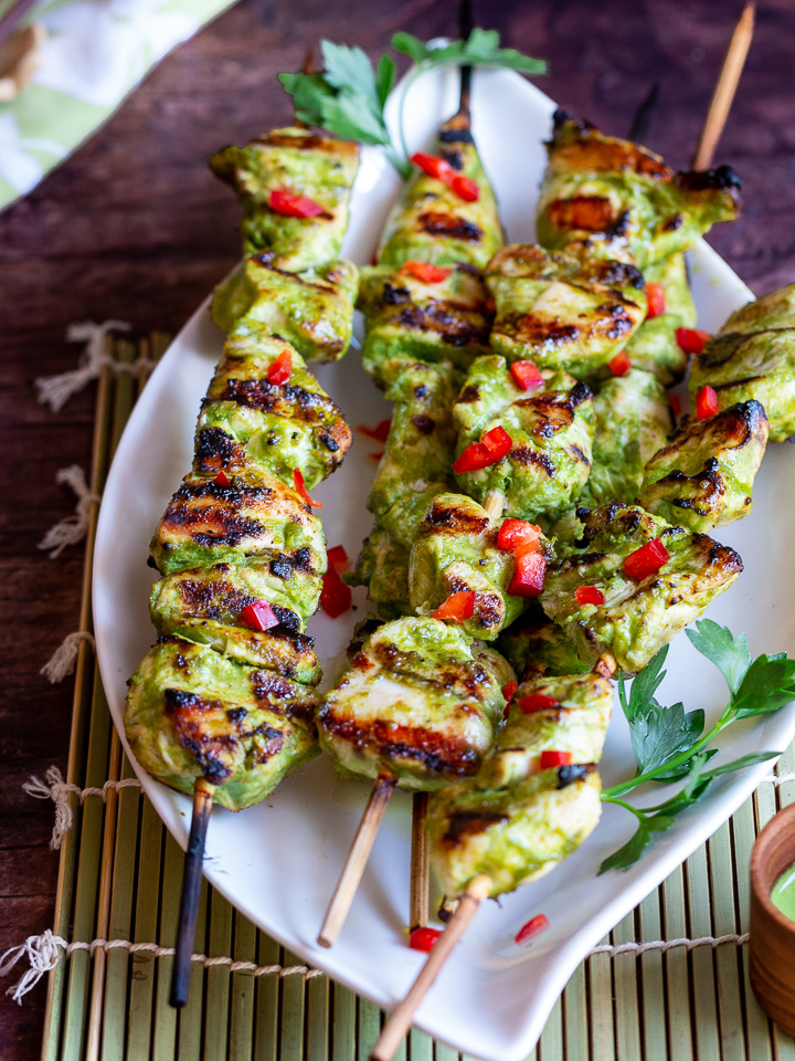 On a green mat with a white plate filled with many chicken skewers and garnished with chili and parsley.