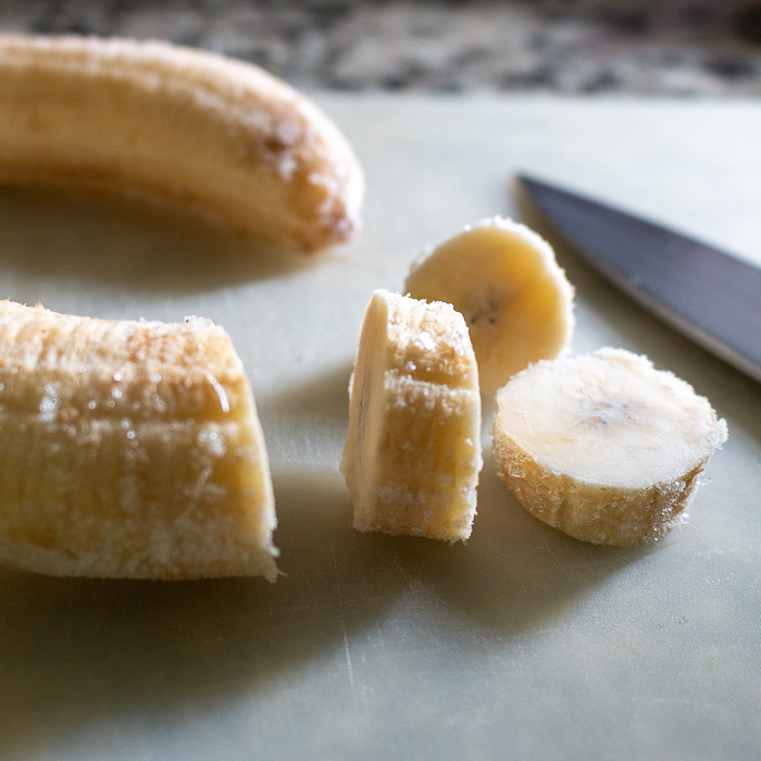 Frozen bananas chopped on a white cutting board with knife on cutting board.