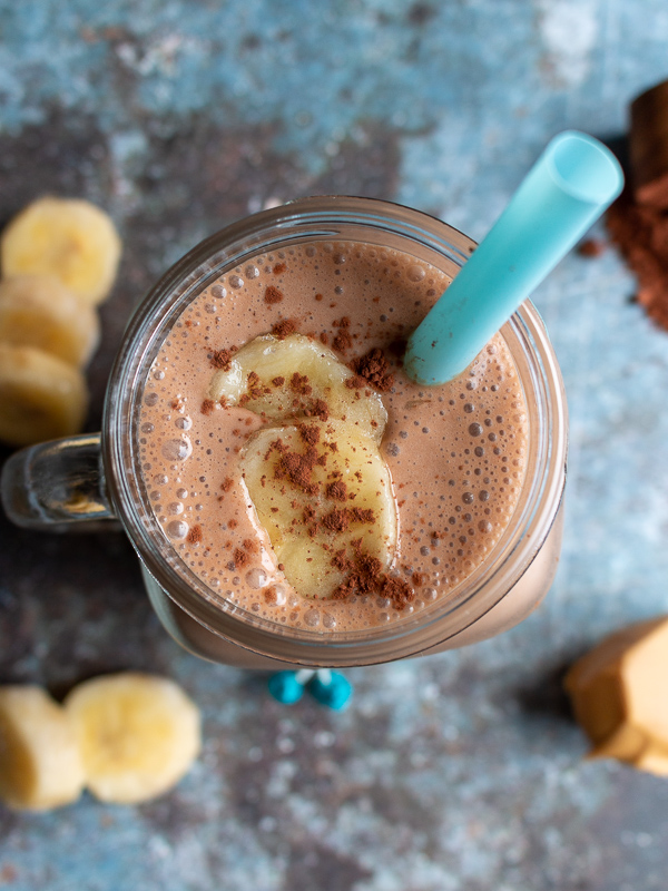 Top Down photo. Creamy rich chocolate peanut butter smoothie topped with bananas and unsweetened chocolate cocoa powder. Surrounded by chopped bananas , peanut butter.