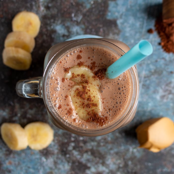 Top Down photo. Creamy rich chocolate peanut butter smoothie topped with bananas and unsweetened chocolate cocoa powder. Surrounded by chopped bananas , peanut butter.