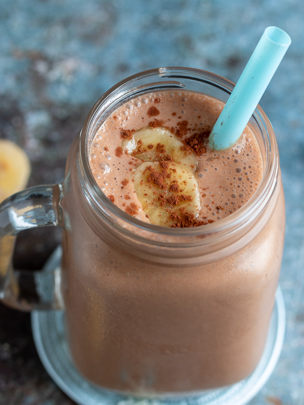 Top Down photo. Creamy rich chocolate peanut butter smoothie topped with bananas and unsweetened chocolate cocoa powder. Surrounded by chopped bananas , peanut butter.