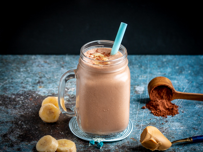 Creamy rich chocolate peanut butter smoothie topped with bananas and unsweetened chocolate cocoa powder. Surrounded by chopped bananas , peanut butter.
