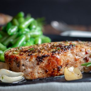 Herb encrusted boneless pork chop on a plate with shallots and cooked green beans in the background.