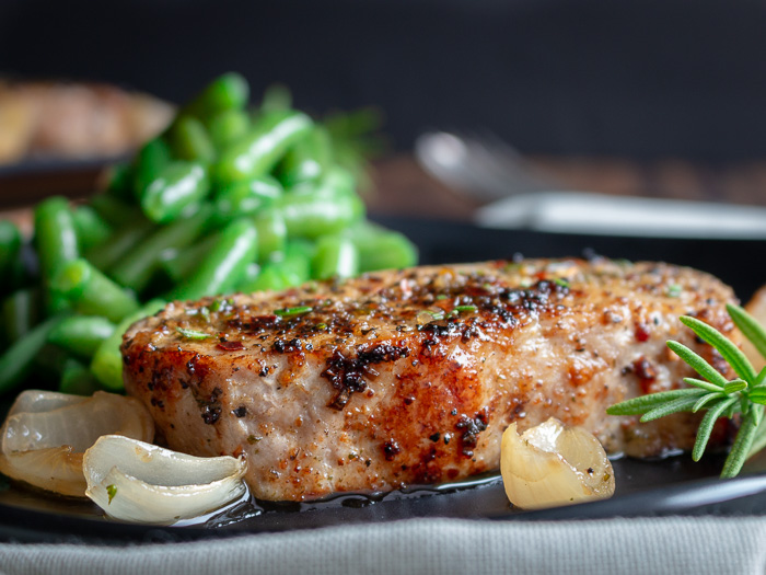 Herb encrusted boneless pork chop on a plate with shallots and cooked green beans in the background.