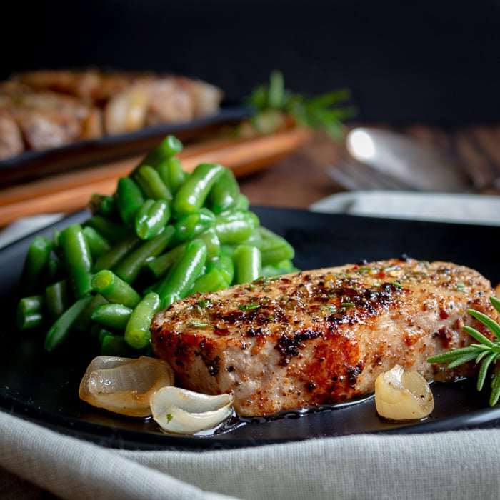 Herb encrusted boneless pork chop on a plate with shallots and cooked green beans and whole tray of pork chops in the background.