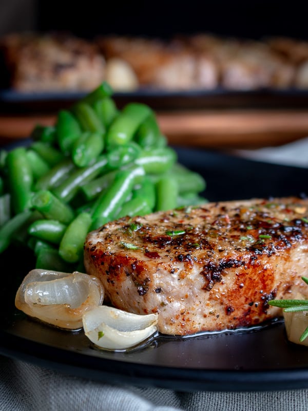 Herb encrusted boneless pork chop on a plate with shallots and cooked green beans in the background.