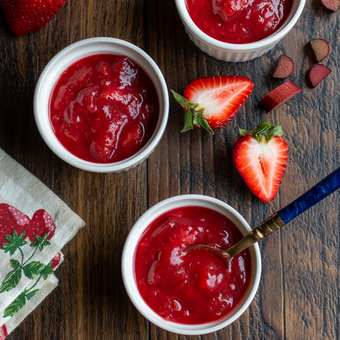 3 bowls of freshly made strawberry jam with a spoon in and fresh strawberries and rhubarb slices around it.