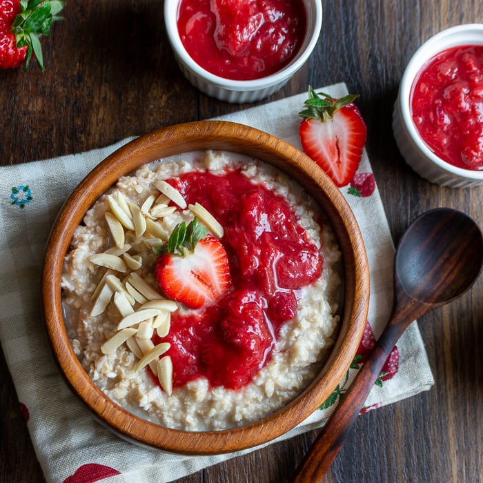 Bowl of oatmeal garnished with strawberry rhubarb jam, almonds and fresh strawberries in a wooden bowl. 