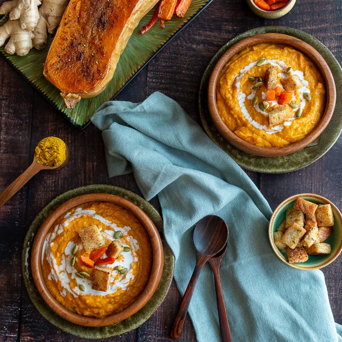 2 bowls of roasted butternut squash soup garnished with pumpkin seeds, coconut milk and homemade croutons.