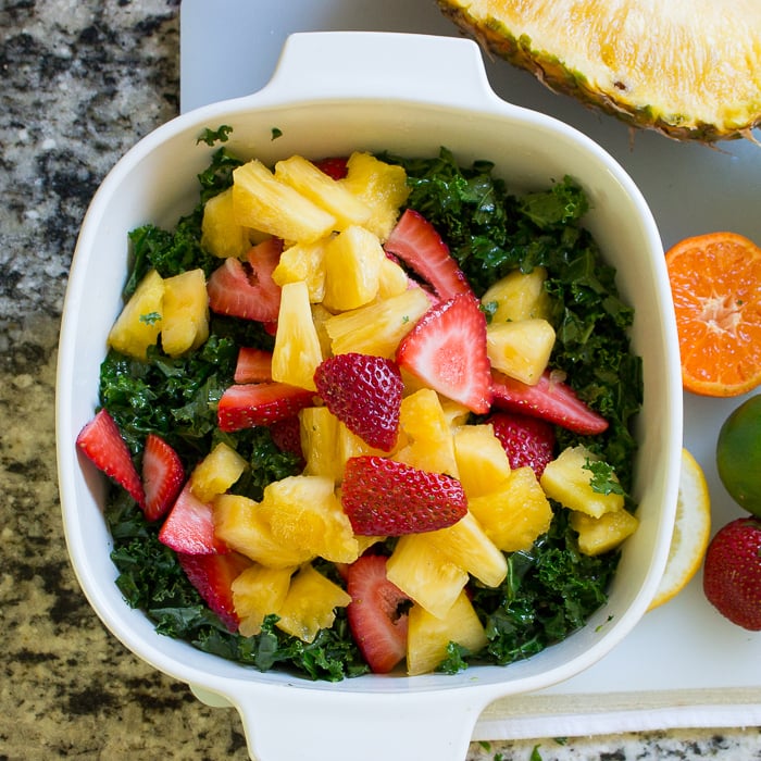 Pineapple and strawberries on top of the kale in a white bowl, ready to be tossed. 