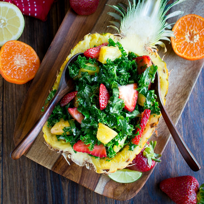 Fruit and kale salat displayed in the pineapple half with 2 cute wooden spoons with fruits along the sides. 